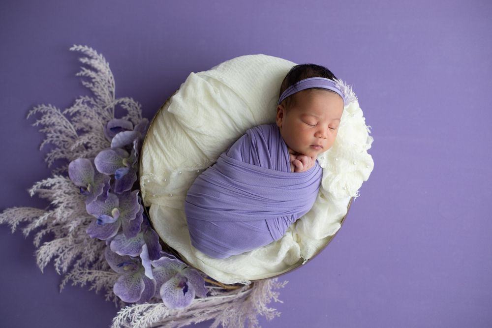 Sleeping newborn wrapped in purple swaddle rests in cream basket with lavender floral decorations on purple backdrop.