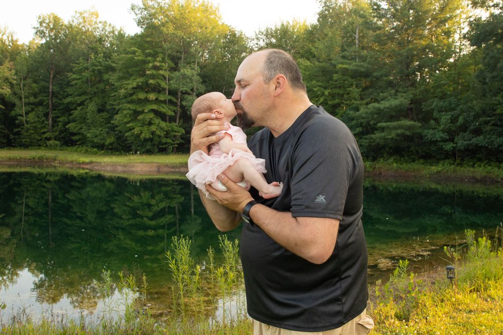 Tender parent-child moment by a pond with autumn foliage in background.