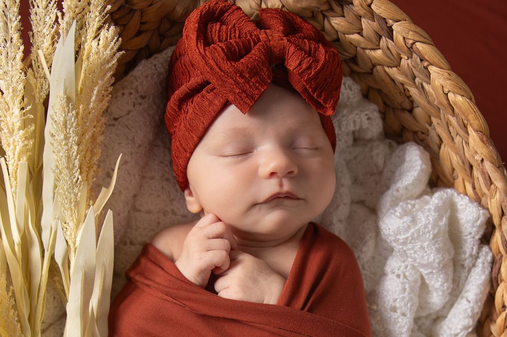 Sleeping newborn wrapped in red fabric with matching bow headband nestled in basket with dried wheat stems.
