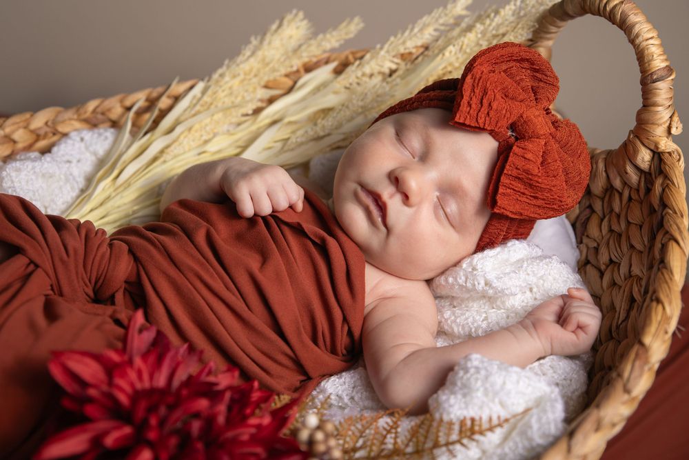 Sleeping newborn in red outfit and bow lying in woven basket with wheat stalks.