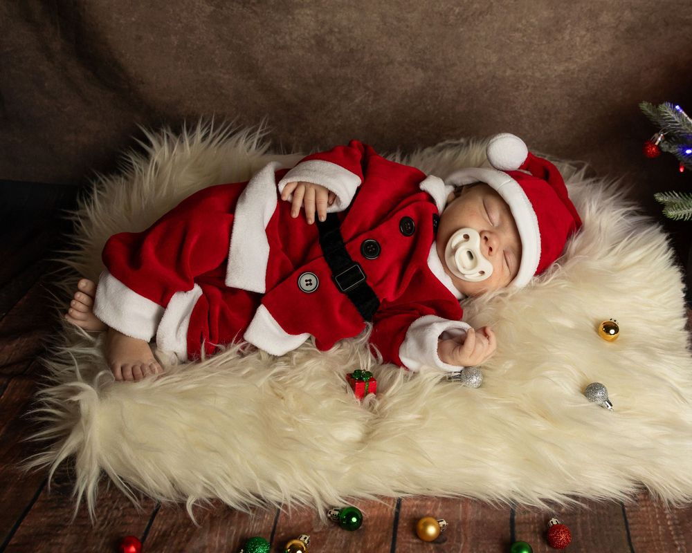 Baby wearing Santa outfit lies on white furry rug with holiday decorations.