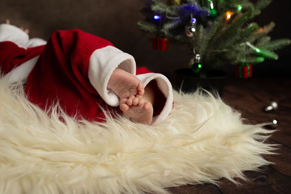 Sleeping baby in Santa outfit on white fur with Christmas tree in background.