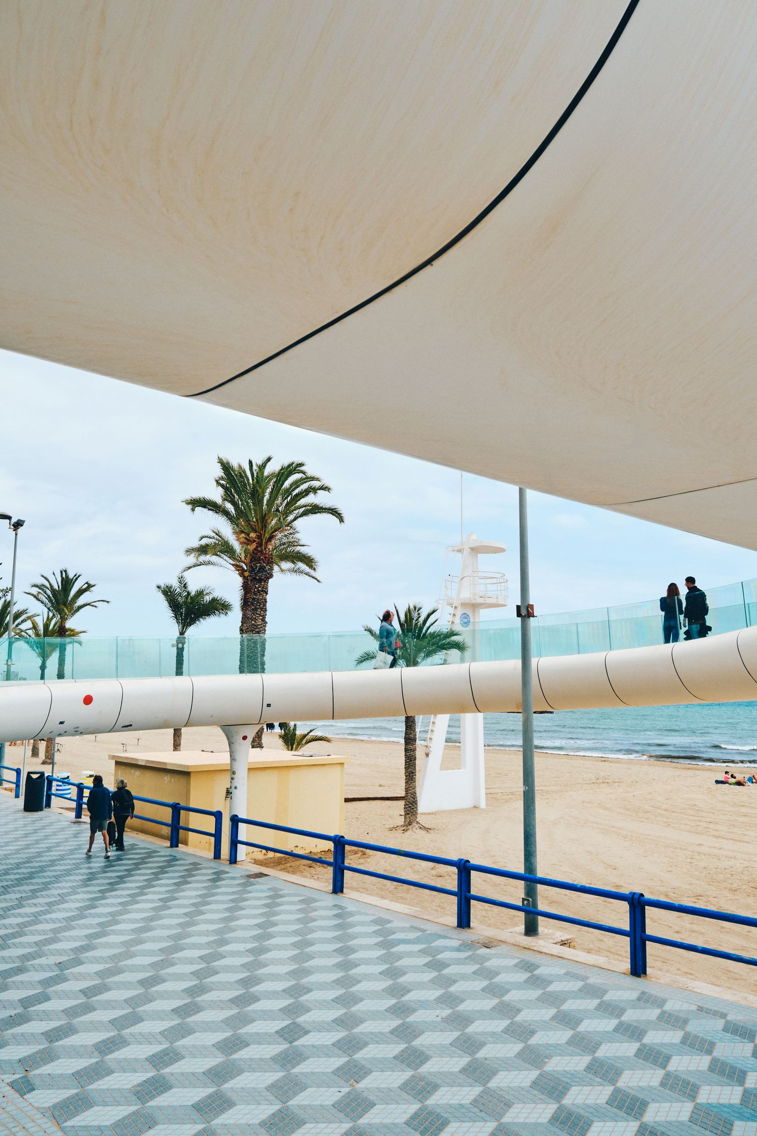 Curved beach promenade with white shade sail, palm trees, and geometric pattern flooring.