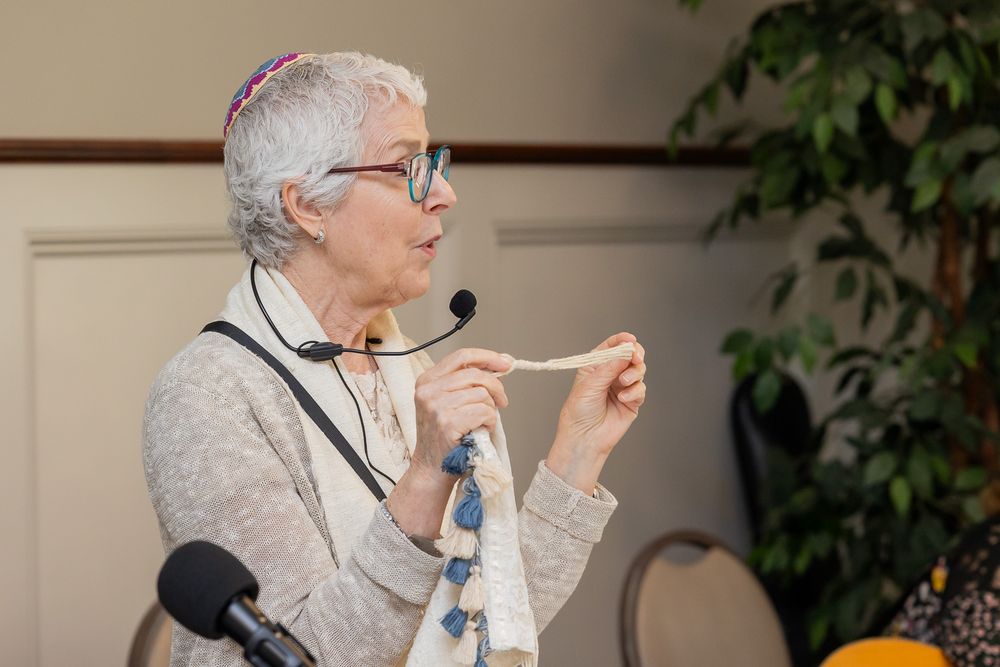 An elderly person in glasses and a gray sweater speaks into a microphone during an event.