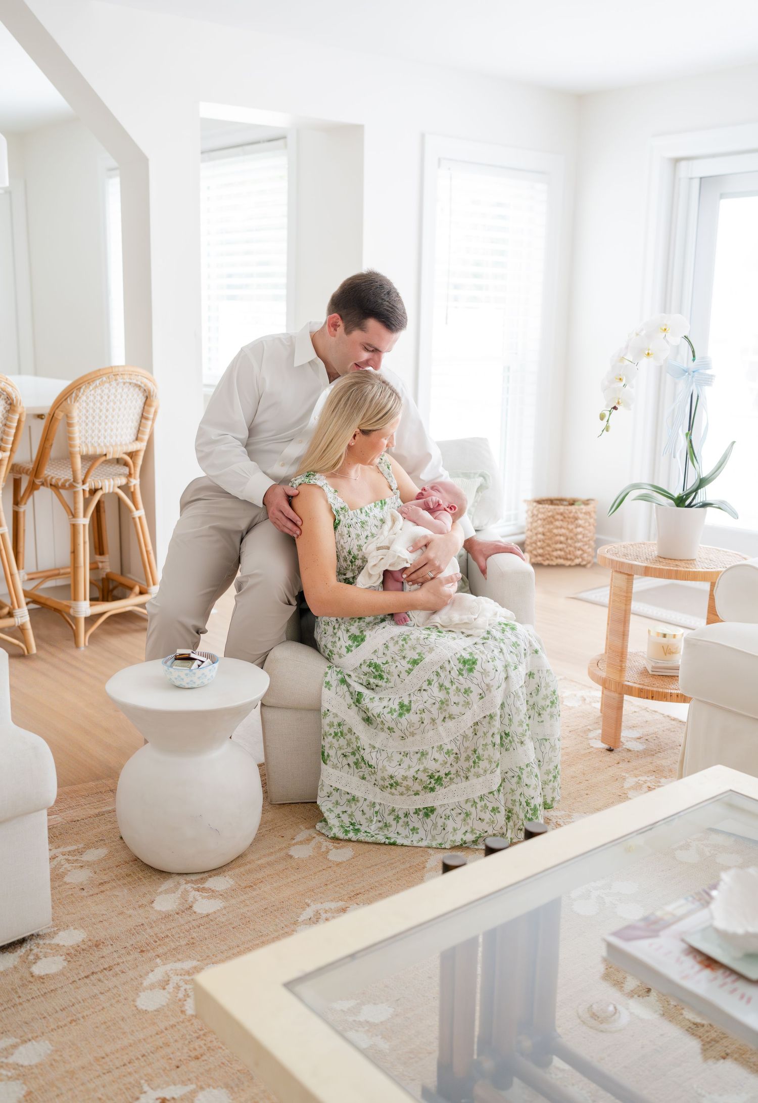 A couple sits on a bed in a bright white room looking adoringly at their newborn baby.