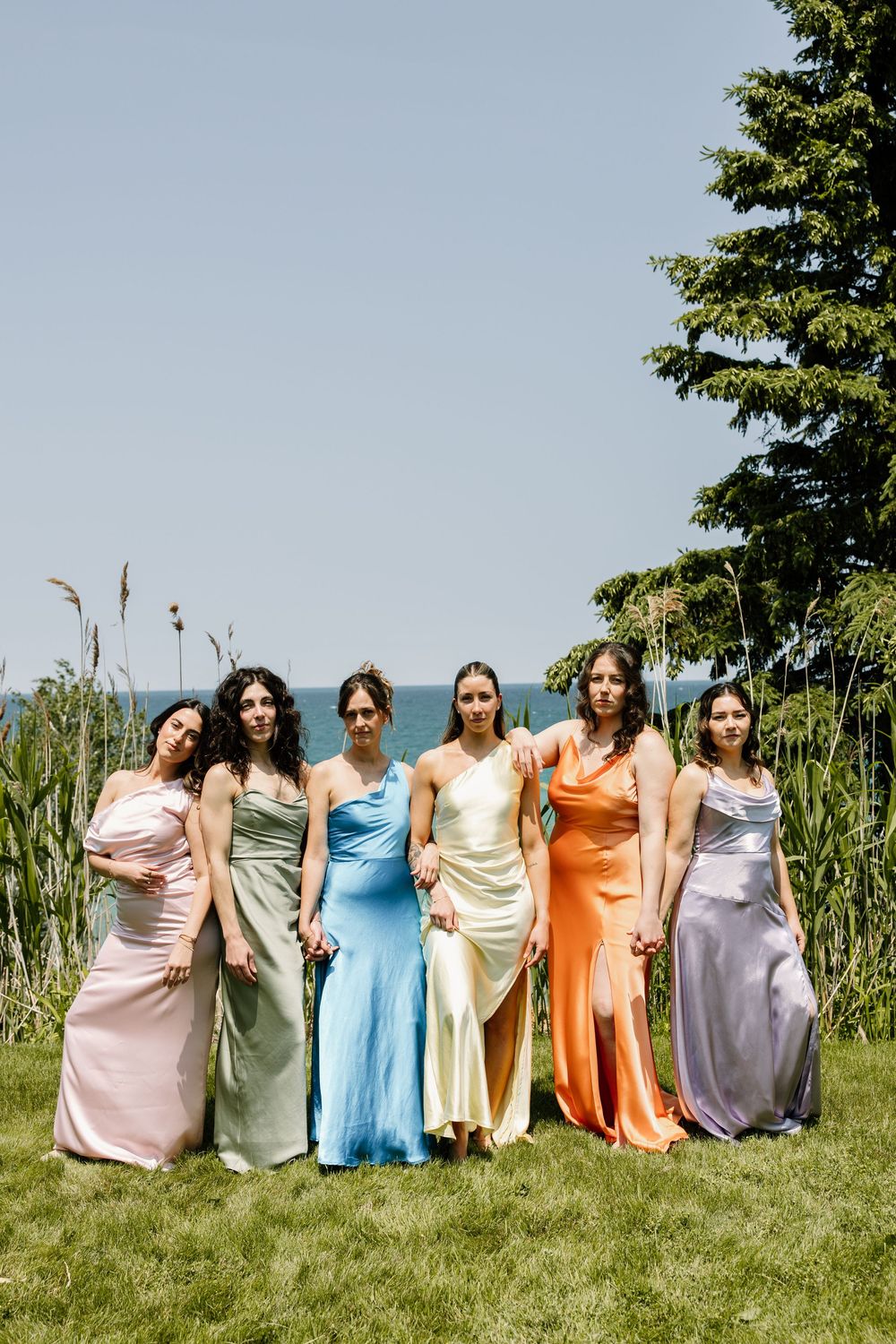 A group in colorful formal dresses stands together on a grassy lawn with trees in the background.