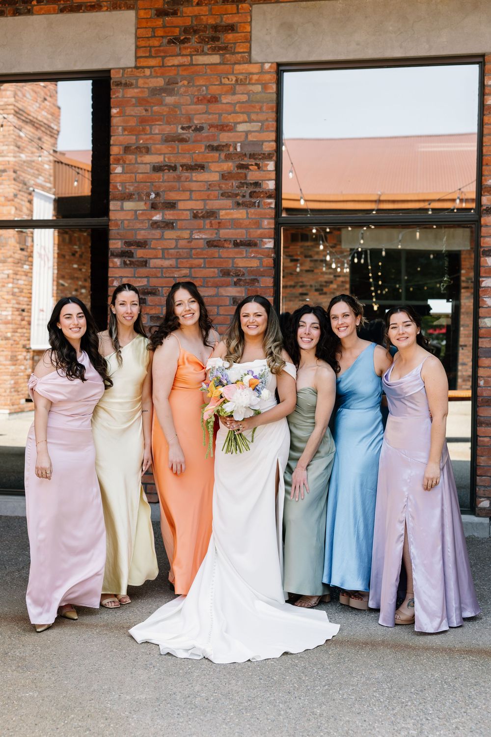 A bridal party poses in pastel-colored dresses outside a brick building with string lights and windows.
