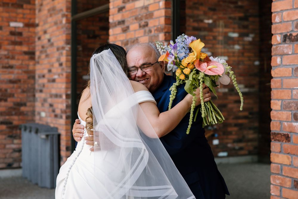 Newlyweds embrace while holding a vibrant wildflower bouquet against rustic brick wall backdrop.