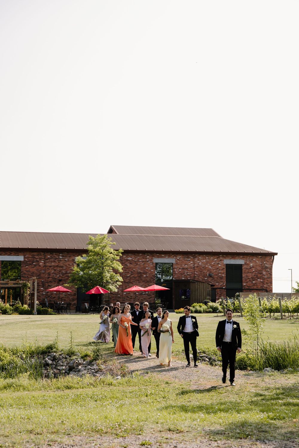 A group of people gather outside a rustic brick venue with red umbrellas and green landscaping on a sunny day.