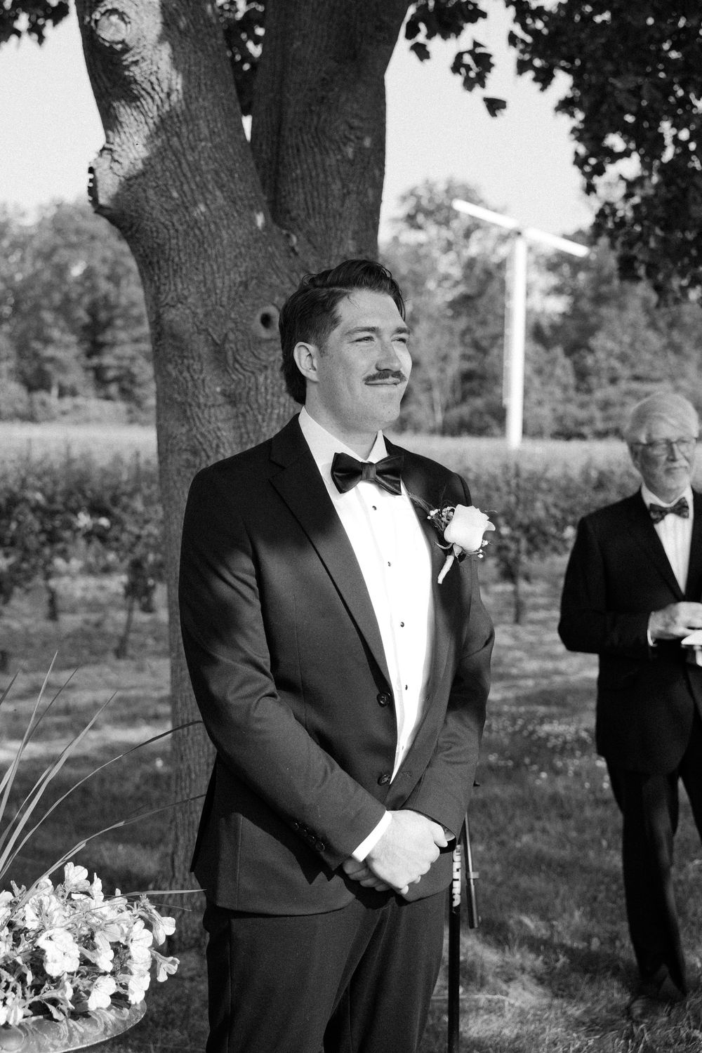 A group in tuxedos share a candid moment outdoors during a formal event beneath tall trees.