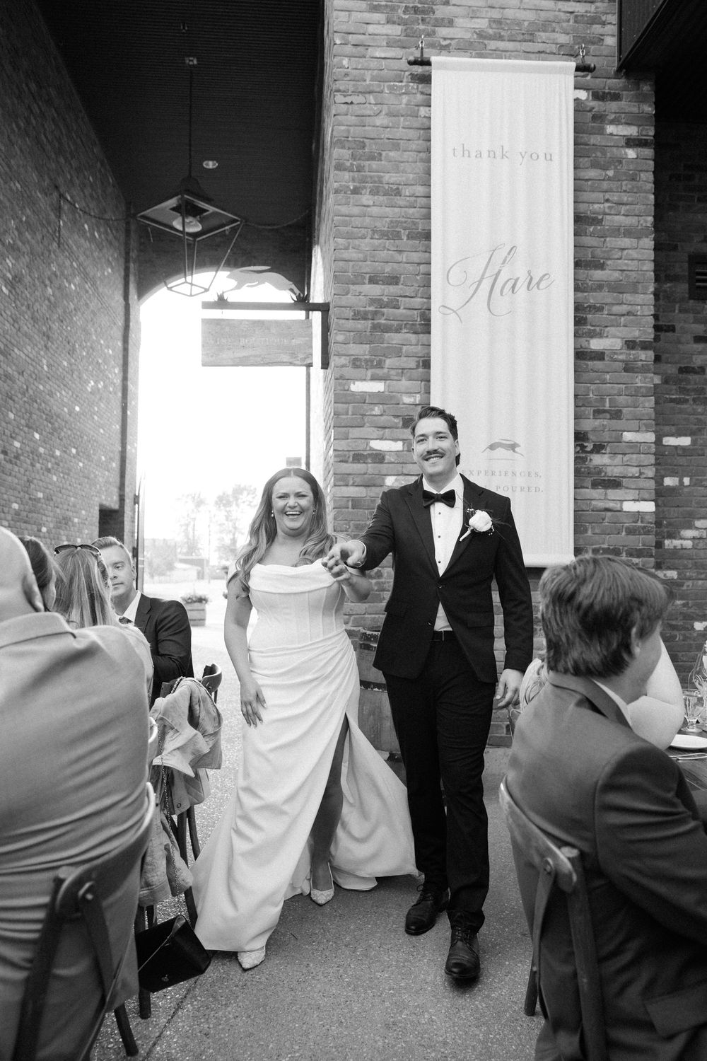 A joyful couple makes their grand entrance at a brick-walled wedding venue in black and white photography.