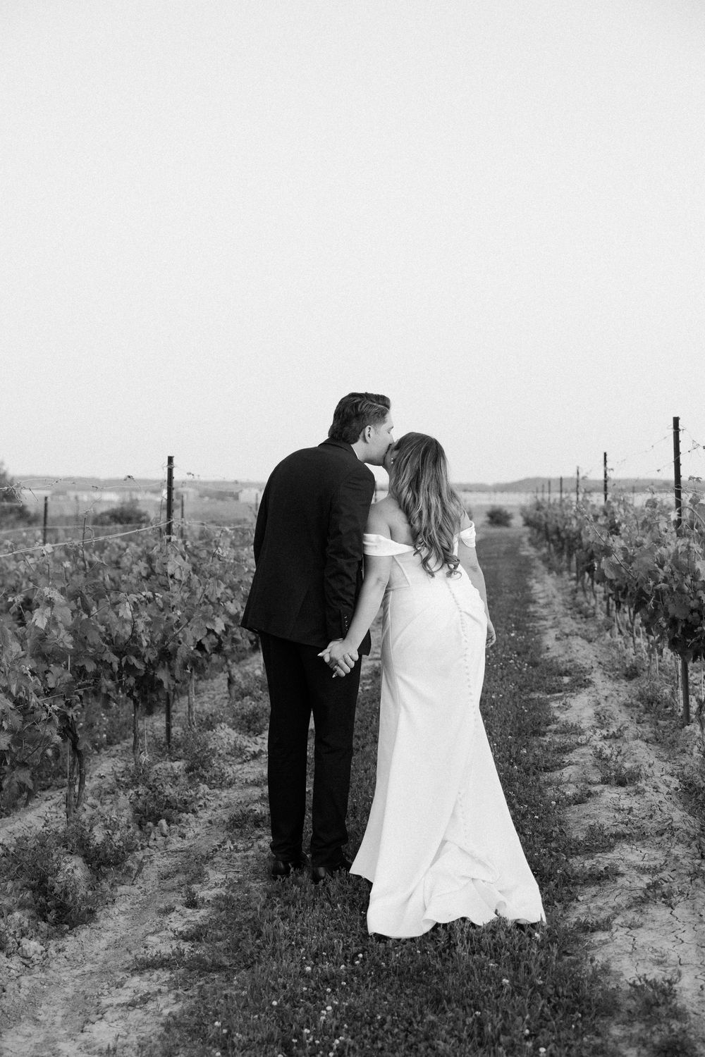 A couple in wedding attire stands between rows of grapevines at a vineyard in black and white photography.