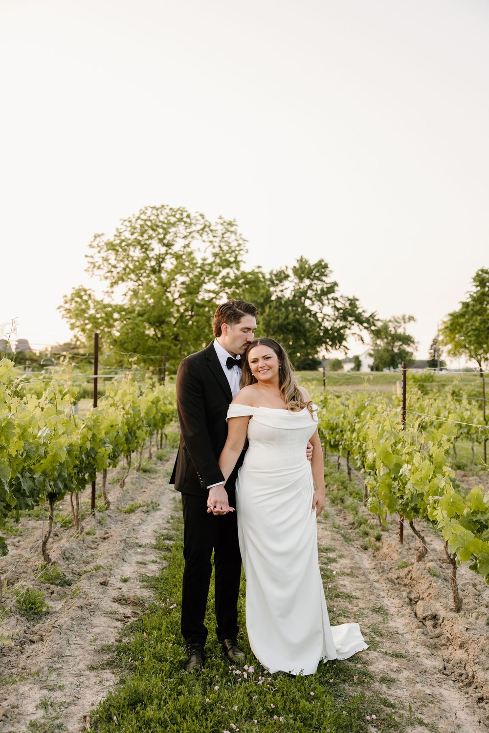 Newlyweds pose together in an outdoor vineyard at sunset, surrounded by rows of grapevines.