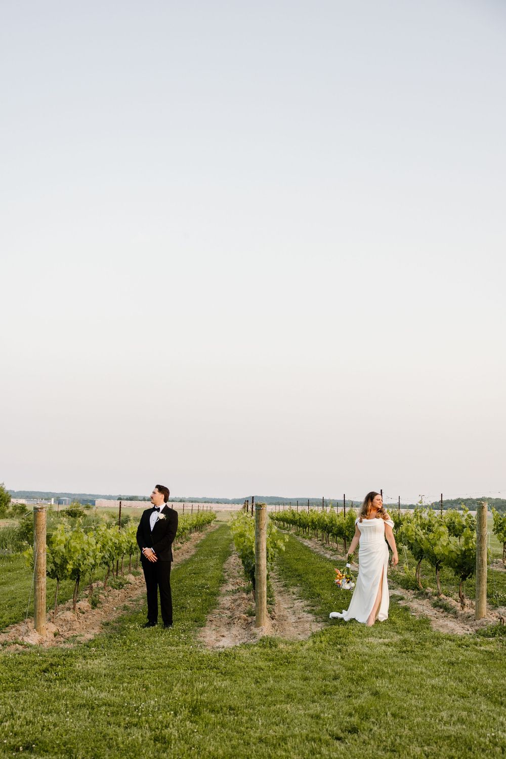 A couple stands apart in a vineyard during a first look photo session.