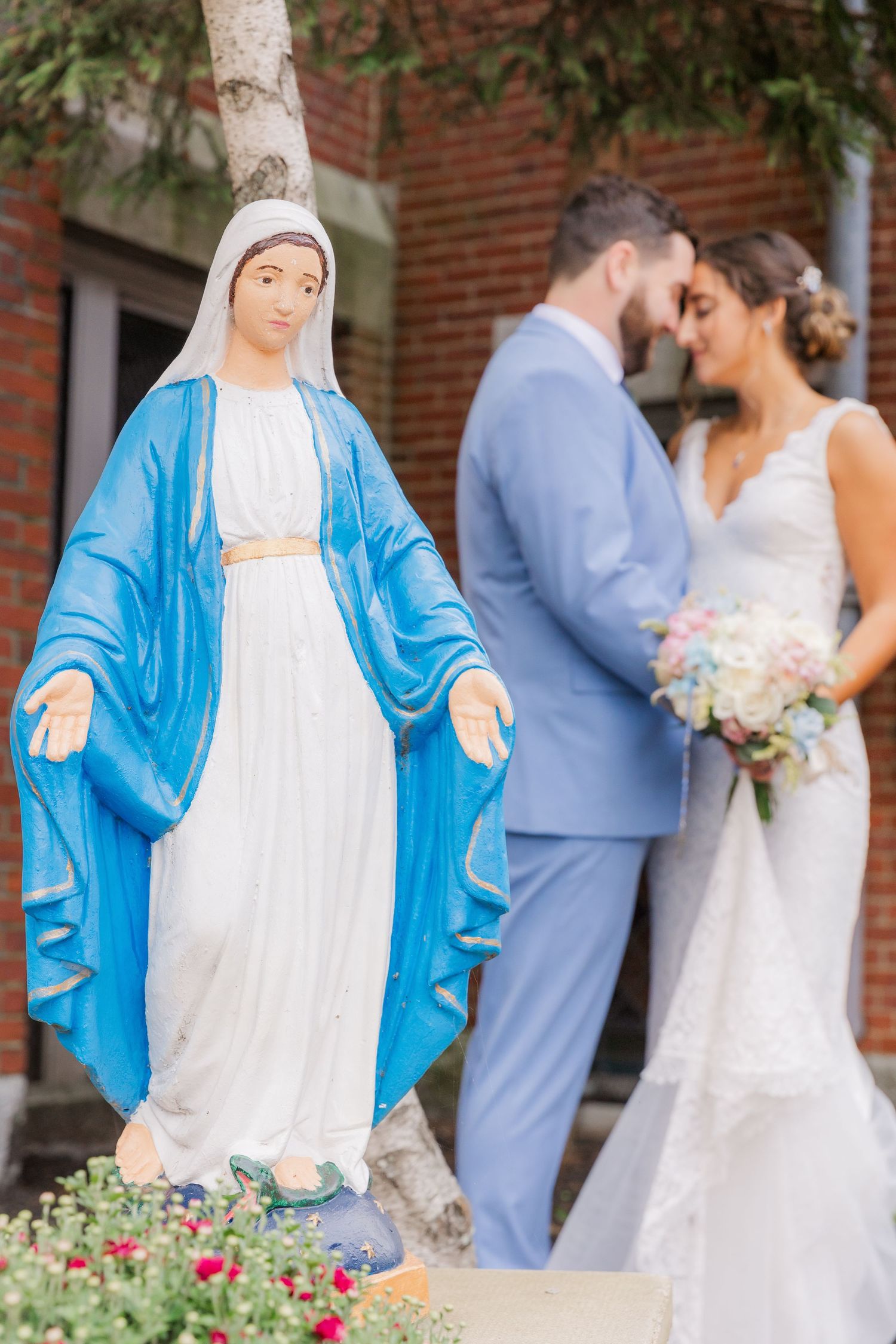Blue and white Madonna statue stands in garden with wedding couple in background.