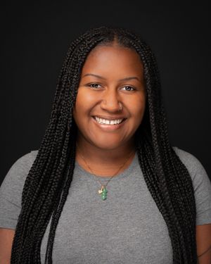 Professional portrait of a person with long braided hair wearing a gray t-shirt and small necklace.
