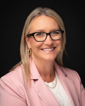 Professional portrait of someone wearing glasses and a pink blazer with white blouse against dark background.