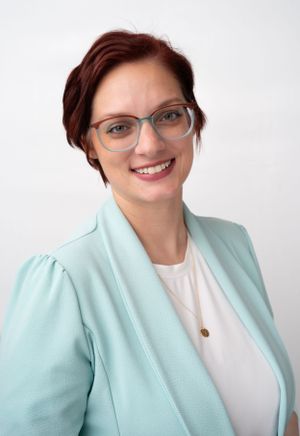 Professional headshot of person in glasses wearing light blue blazer and white top against white background.