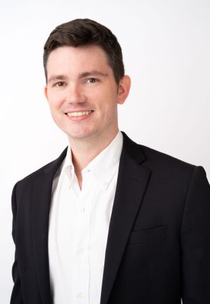 Professional headshot of person in black suit and white shirt against white backdrop.