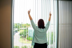 Person raising arms triumphantly while facing large office windows with city view.
