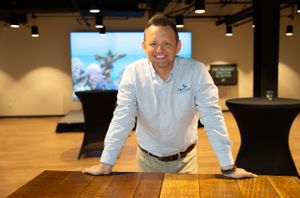 Professional in white shirt standing confidently at wooden table in open office space.