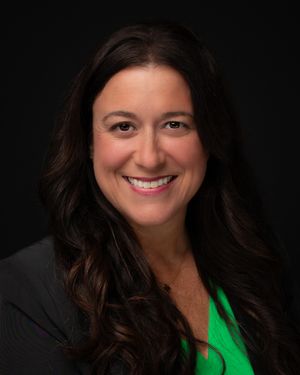 Professional headshot of a person in a dark blazer and bright green top against a black background.