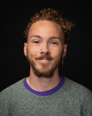 A person with curly ginger hair and beard wearing a gray sweater with purple collar against a dark background.