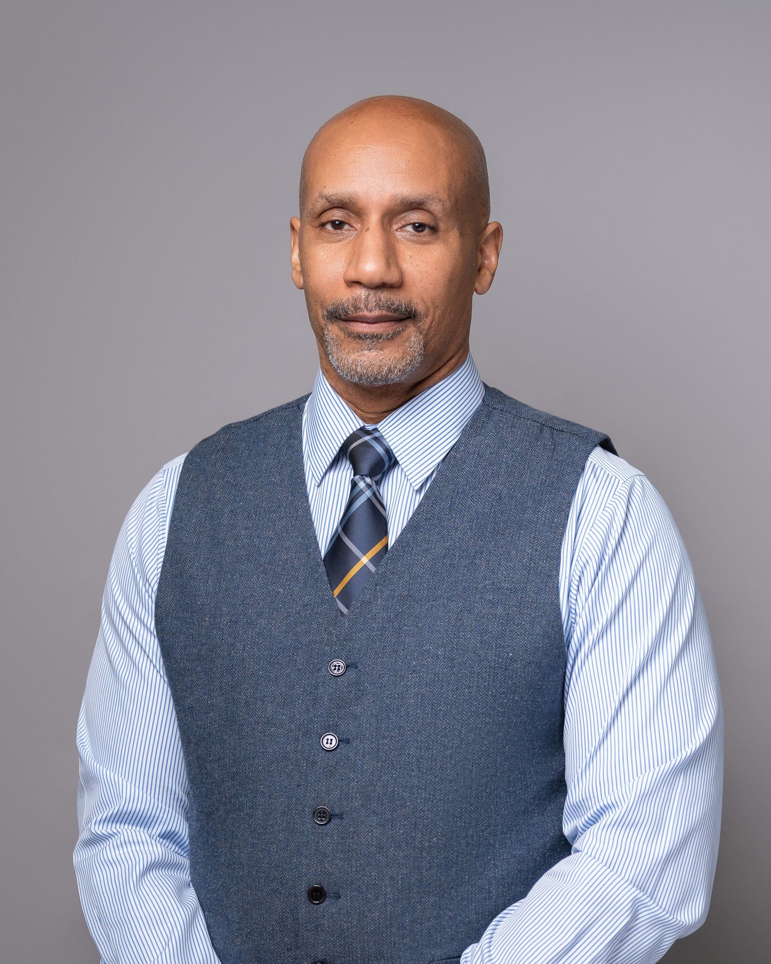 Professional headshot of person wearing grey vest and striped shirt with blue tie against neutral background.