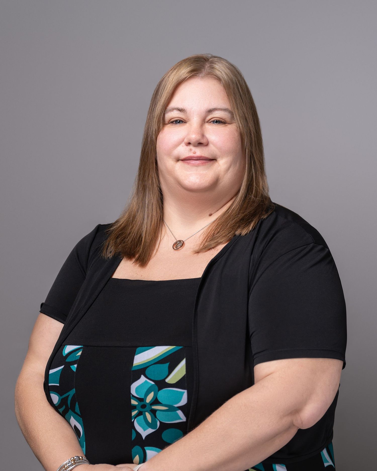 Professional headshot of person in black cardigan and floral dress against grey background.