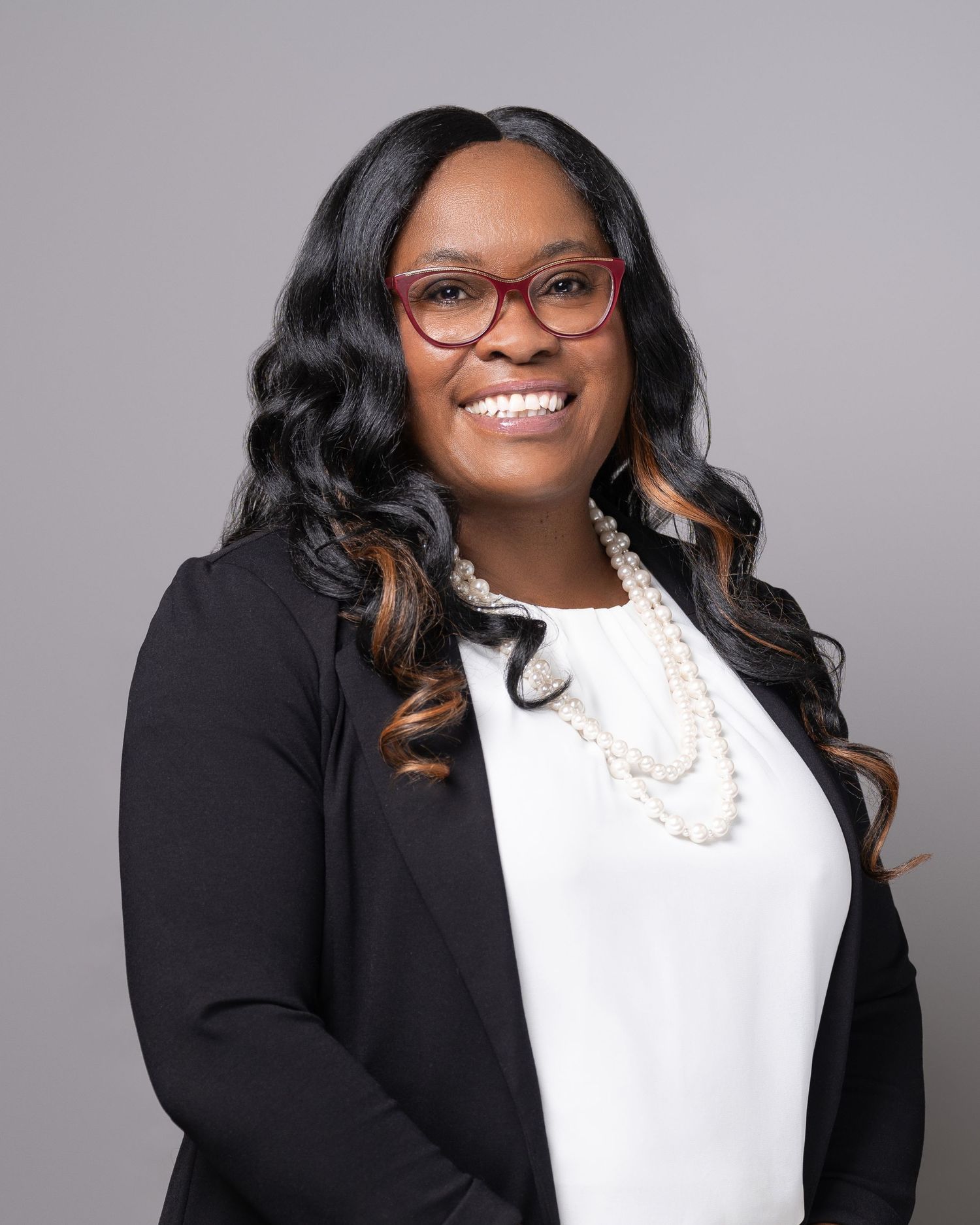 Professional headshot wearing black blazer with pearl necklace and glasses.