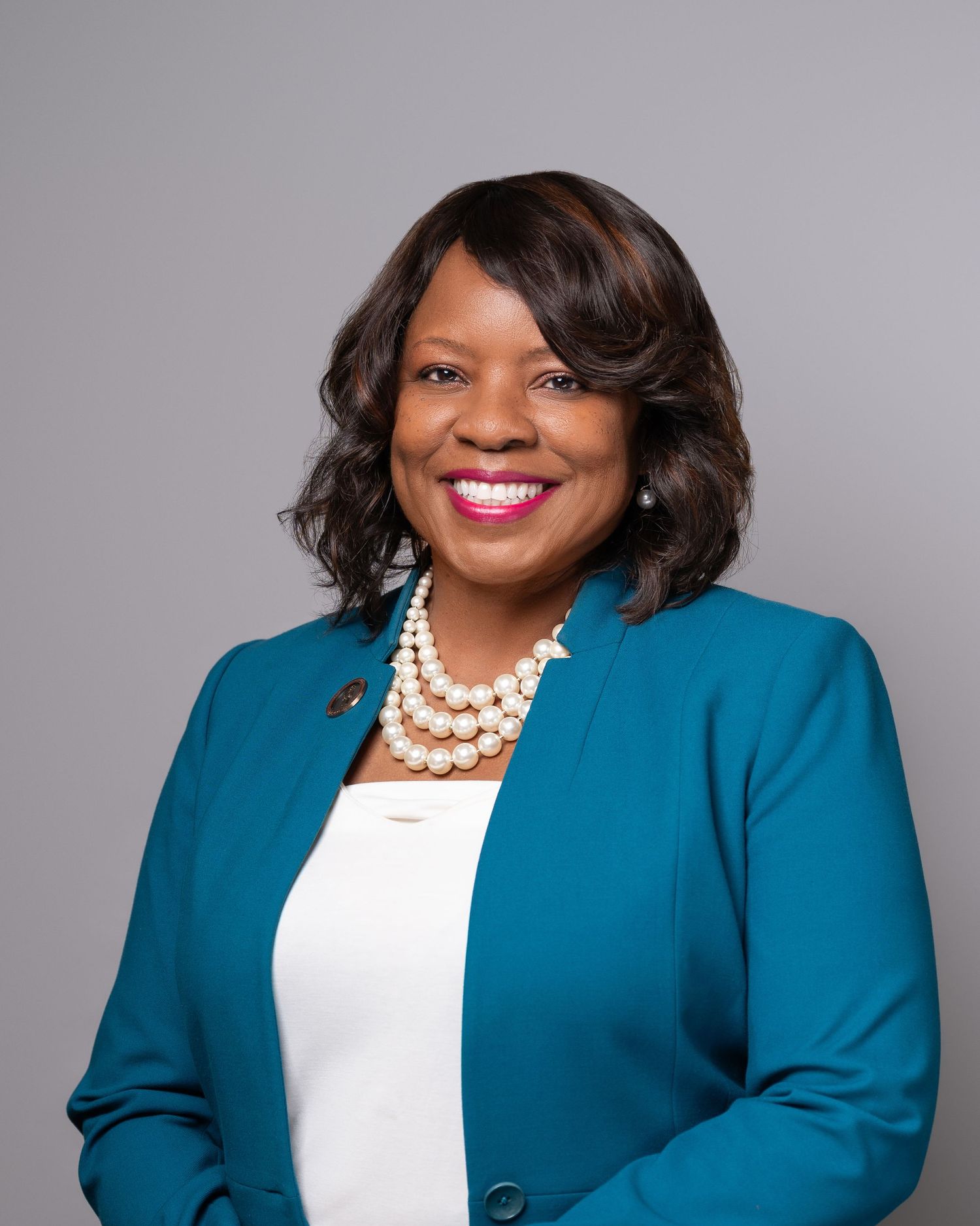 Professional portrait in teal blazer with pearl necklace against gray background.