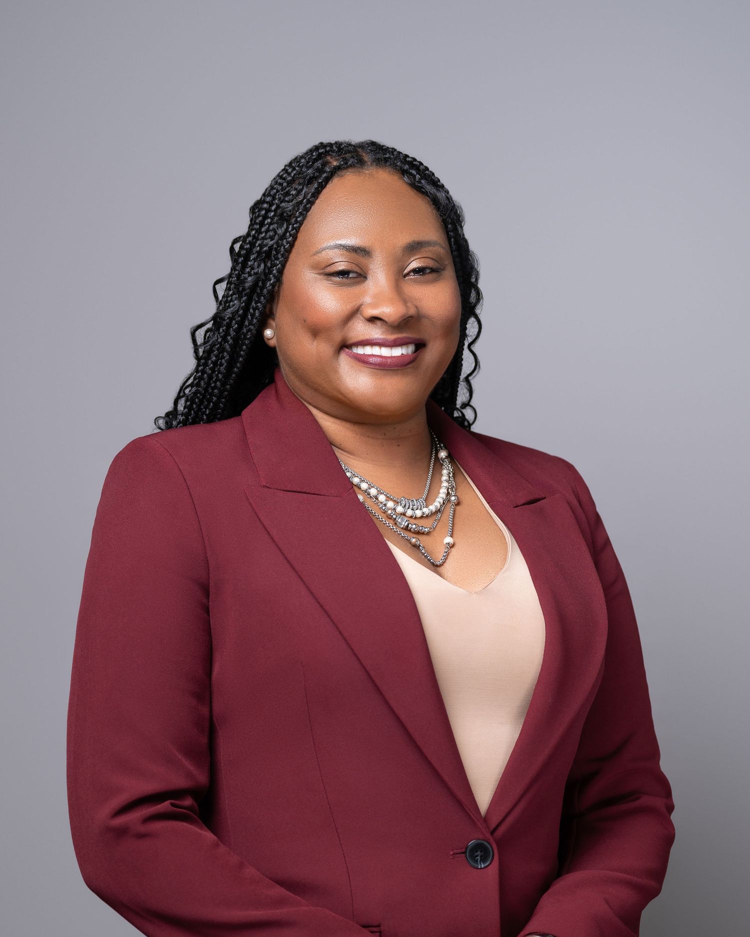 Professional headshot in burgundy blazer with braided hair and pearl necklace.