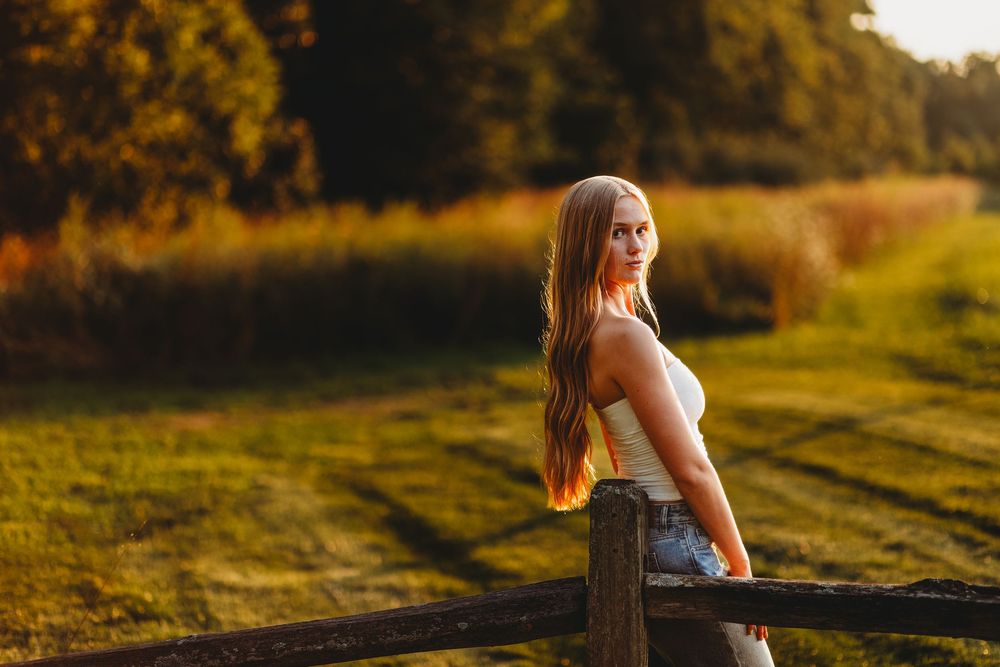 Young woman with long hair in white top leaning on wooden fence at golden hour in a lush green field.