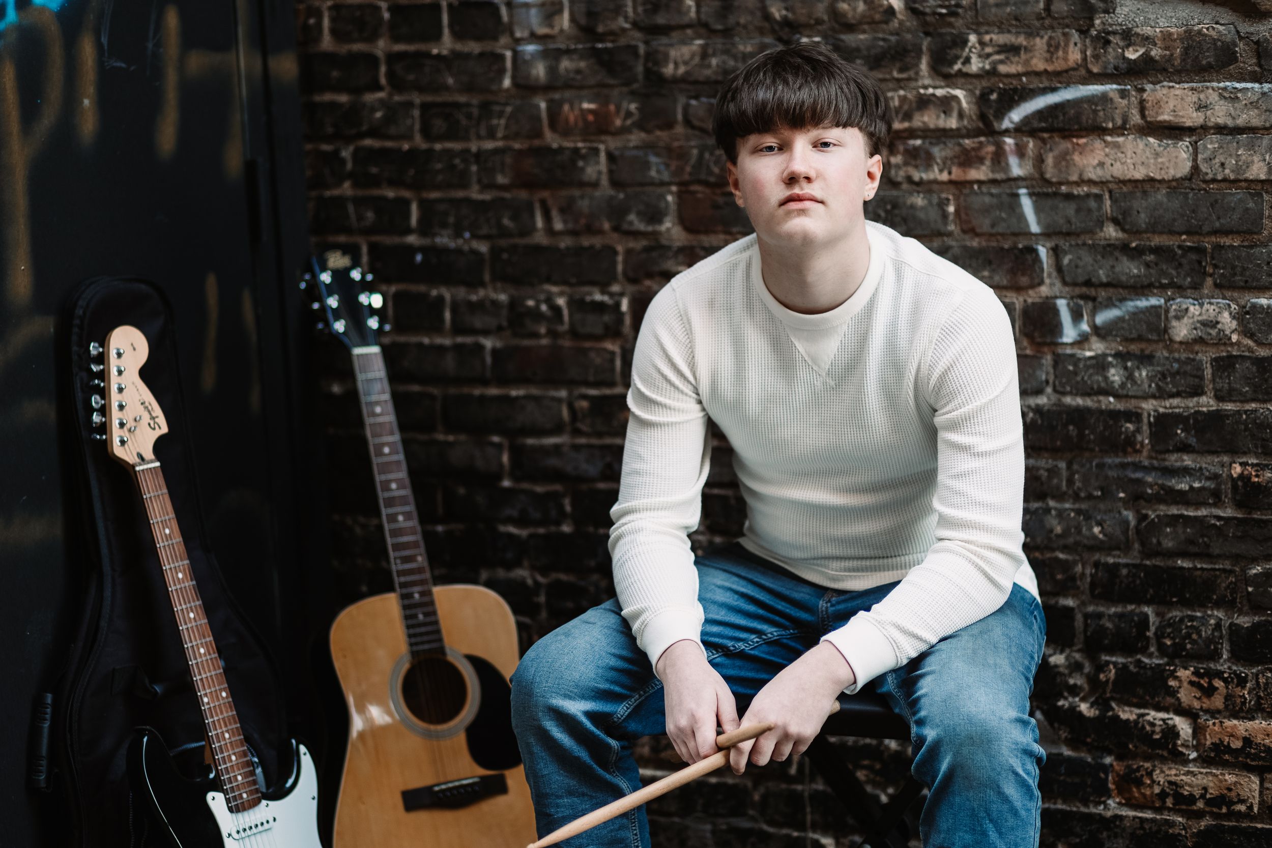 Young musician sits against brick wall with acoustic and electric guitars, holding drumsticks, wearing white sweater and jeans.