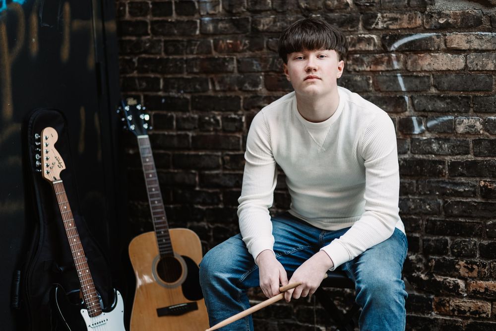 Young musician sits against brick wall with acoustic and electric guitars, holding drumsticks, wearing white sweater and jeans.