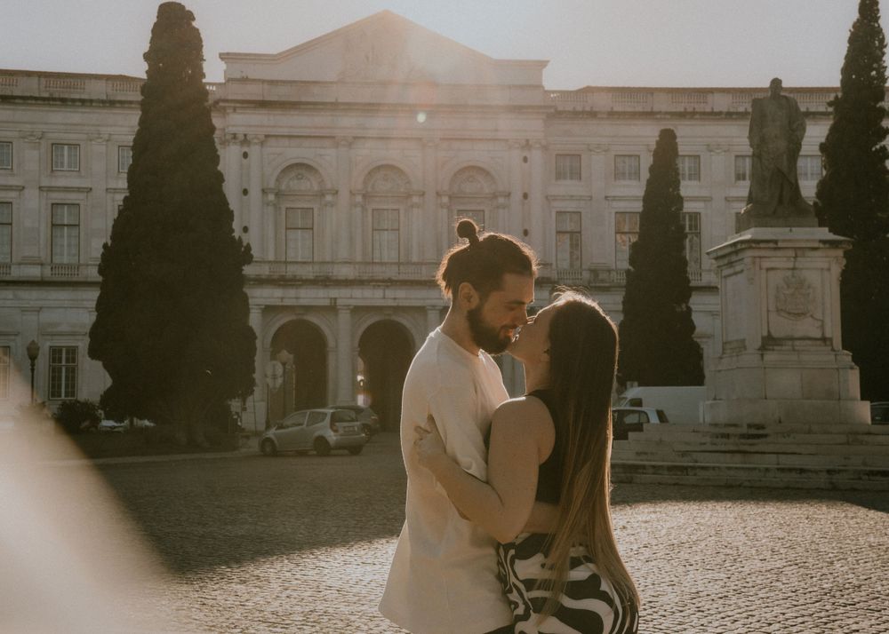 Romantic couple embracing in courtyard with historic building and cypress trees in background.