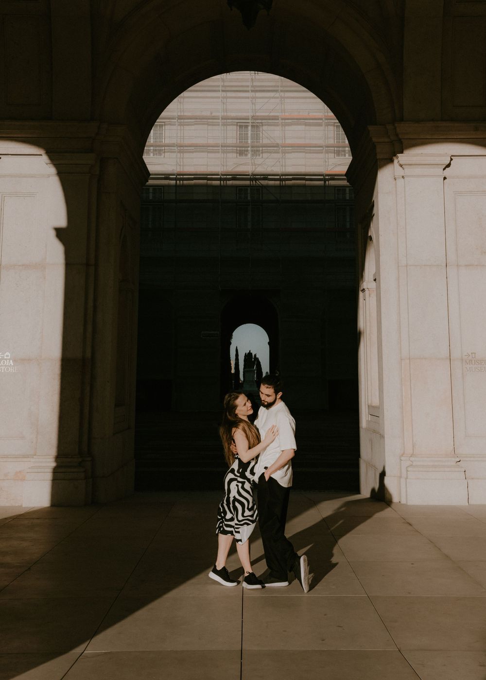 A couple stands together in dramatic lighting under a series of stone archways.