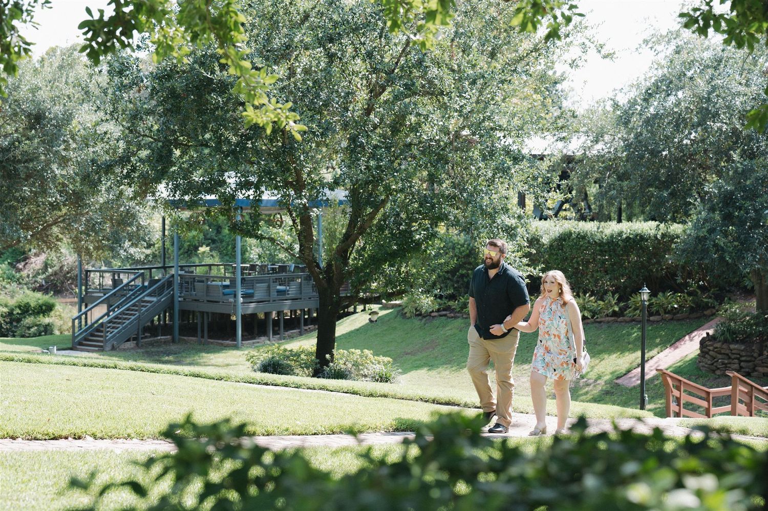 A couple walks together on a sunny path in a lush park setting with a playground in the background.