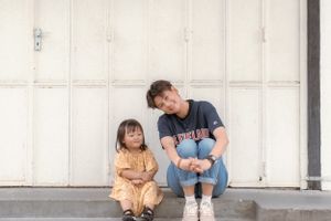 Two figures sitting together against a white wall in casual attire.