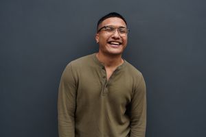 Smiling person wearing an olive green henley shirt stands against a dark grey wall.