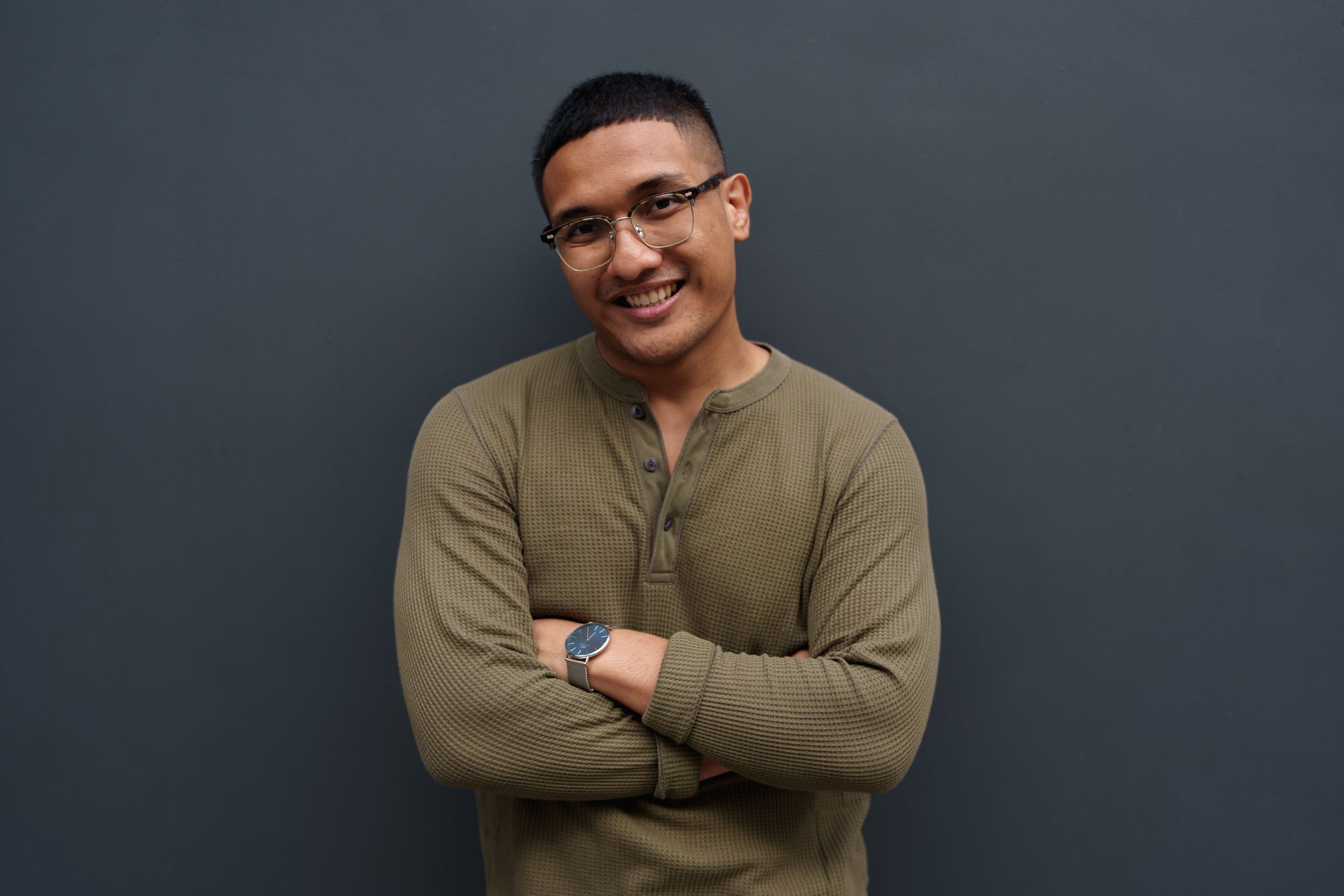 A casually dressed person in an olive henley shirt smiles while standing against a dark gray wall with crossed arms.