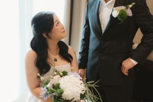 Wedding couple dressed in formal attire with white floral bouquet and boutonniere standing by window indoors.