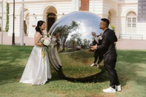 Two people hold a large reflective mirrored orb during a wedding photoshoot on a lawn near a historic building.