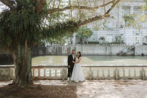 A couple in wedding attire stands near a tree by a waterfront stone balustrade on a romantic overcast day.