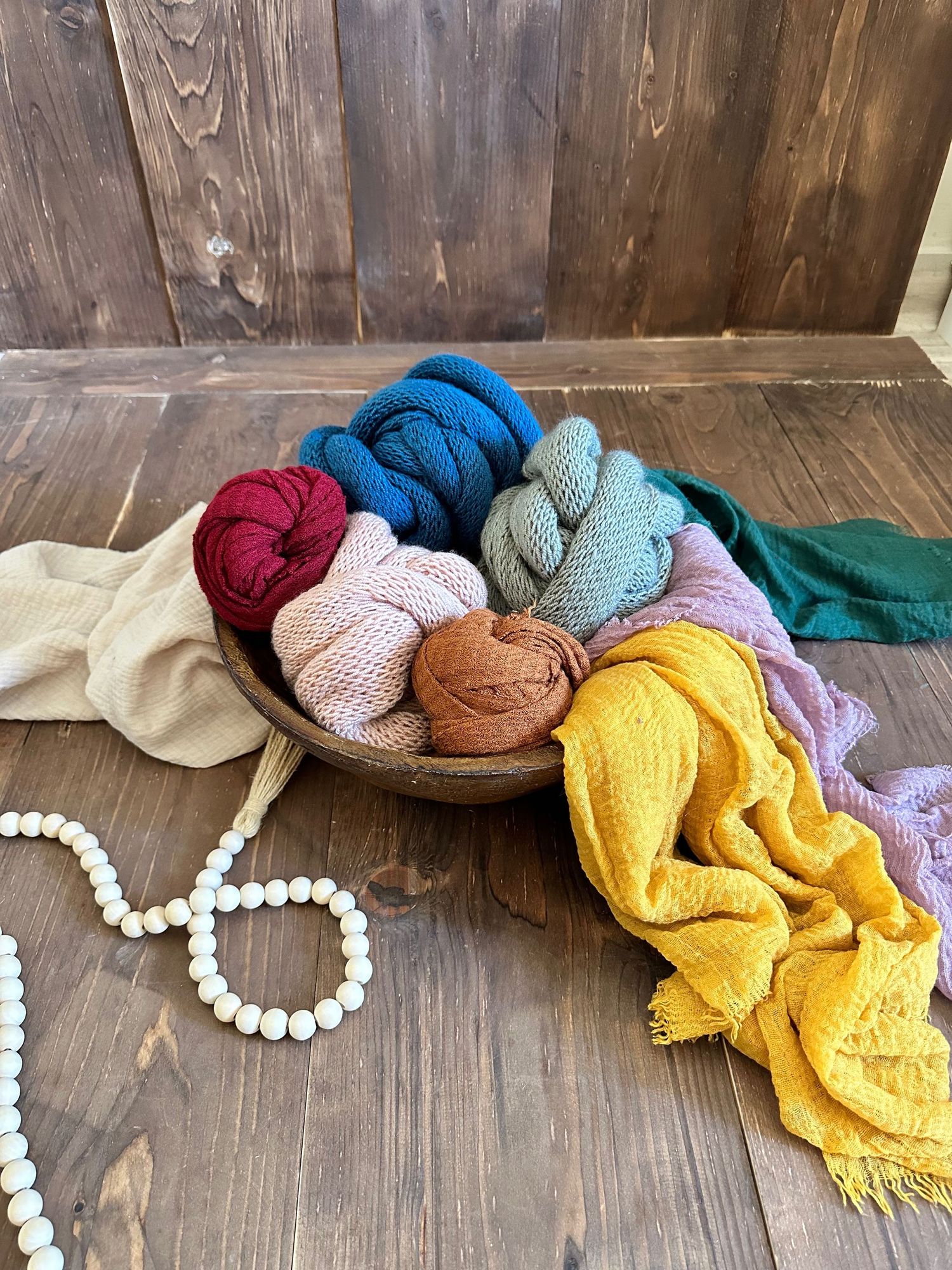 Colorful knit scarves and accessories arranged in a wooden bowl with decorative beaded garland on wooden surface.