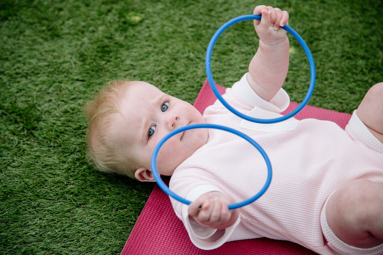 Baby lying on pink yoga mat playing with blue exercise rings on artificial grass.