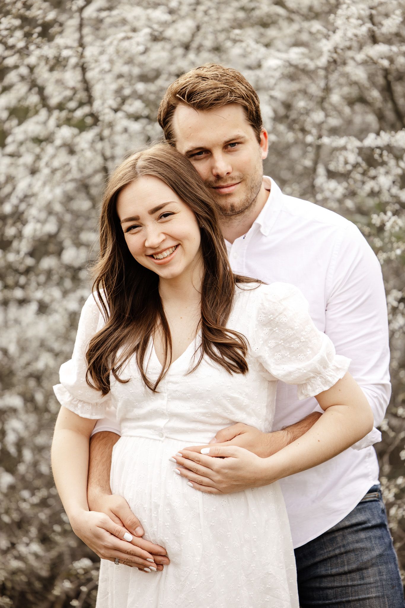 A couple in white outfits embraces during a maternity photoshoot with blooming trees in the background.