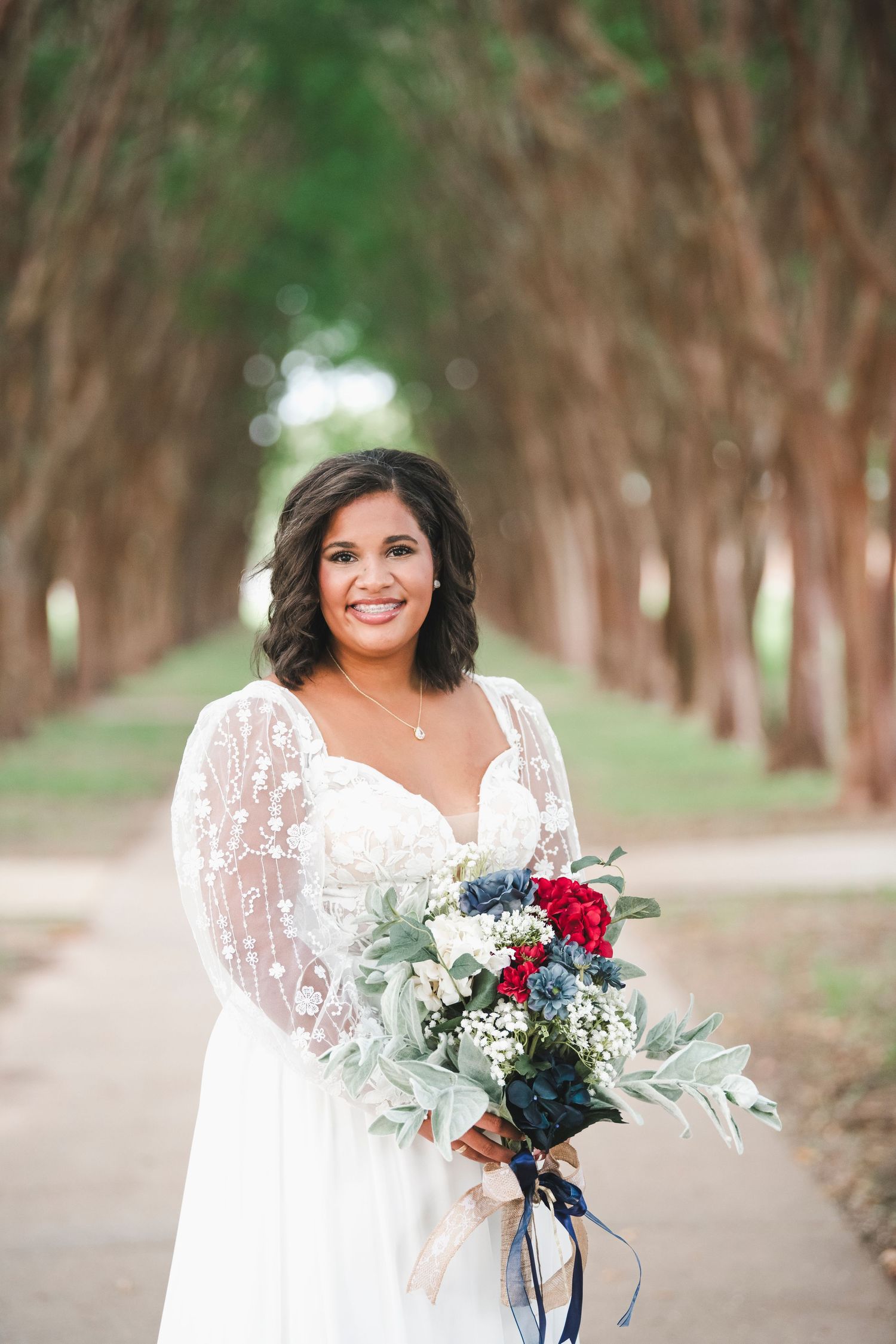 Joyful bride holds colorful bouquet while standing in tree-lined pathway.