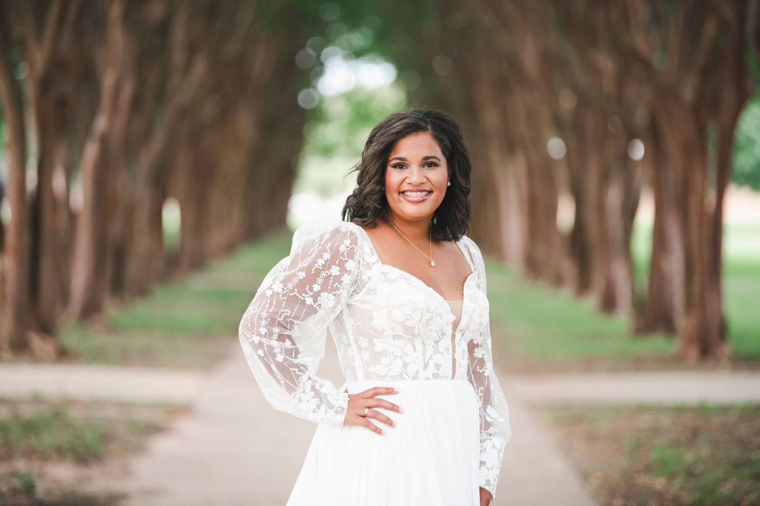Bride in lace sleeve wedding dress poses in tree-lined outdoor pathway.