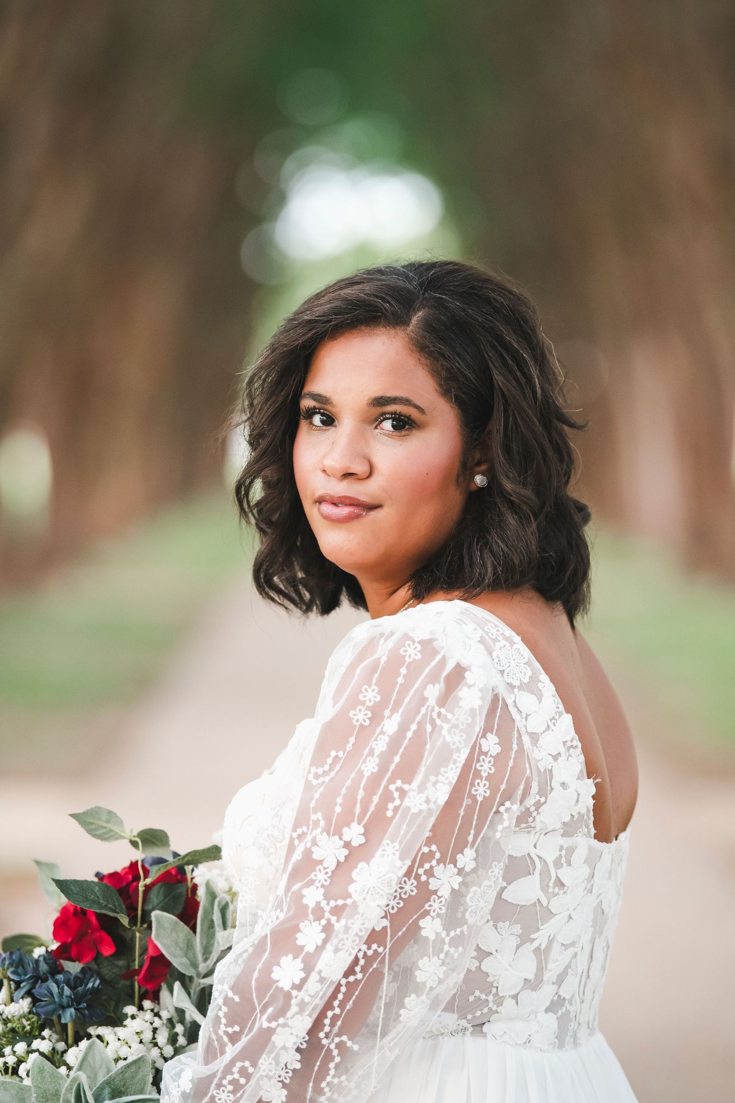 Elegant bridal portrait showcasing lace details and red rose bouquet against tree-lined backdrop.
